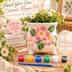 Può includere: Un vaso da fiori in ceramica bianca con un motivo floreale rosa in rilievo e tre piccoli piedini. Il vaso è accompagnato da un set di sei piccoli barattoli di vernice nei colori rosso, giallo, blu, verde, bianco e nero, e un pennello blu.