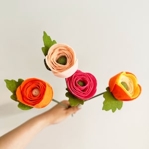 Pu&ograve; includere: Una mano che tiene quattro fiori di feltro nei toni dell'arancione, del rosa e del pesca. Ogni fiore ha uno stelo e foglie verdi. I fiori sono in piena fioritura, con una costruzione dettagliata dei petali.