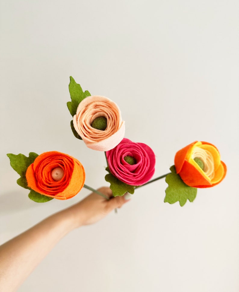 Pu&ograve; includere: Una mano che tiene quattro fiori di feltro nei toni dell'arancione, del rosa e del pesca. Ogni fiore ha uno stelo e foglie verdi. I fiori sono in piena fioritura, con una costruzione dettagliata dei petali.