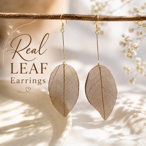 May include: A pair of gold-toned drop earrings featuring delicate, skeletonized brown leaves suspended from thin metal wires. The text "Real LEAF Earrings" is printed in brown on the left side of the image, accompanied by a small heart icon.