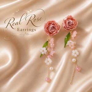 May include: A pair of earrings featuring pink rose-shaped tops with dangling gold chains, green leaves, small white and pink flower accents, and round beads. The text Real Rose Earrings is visible in the upper left corner of the image.