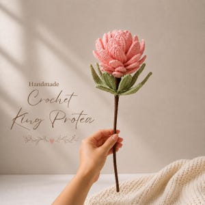 May include: A crochet King Protea flower featuring layered, soft pink petals and sage green leaves attached to a long, thin brown stem. The text "Handmade Crochet King Protea" is printed to the left of the flower with a small heart icon.