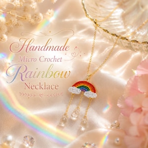 May include: A gold-tone chain necklace featuring a micro-crochet rainbow pendant with white clouds at each end. Three delicate gold chains hang from the rainbow, each ending in a clear, teardrop-shaped bead. Text reads: Handmade Micro Crochet Rainbow Necklace.