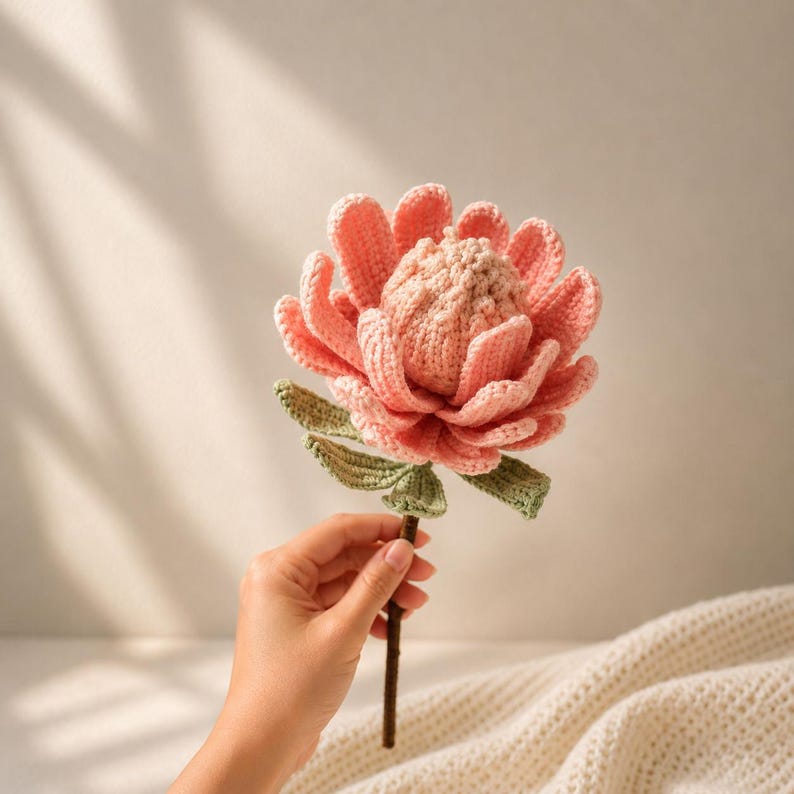 May include: A handcrafted crochet flower featuring a textured, light pink central dome surrounded by layered, soft pink petals. The flower includes small green leaves attached to a thin, brown stem, creating a detailed botanical representation.
