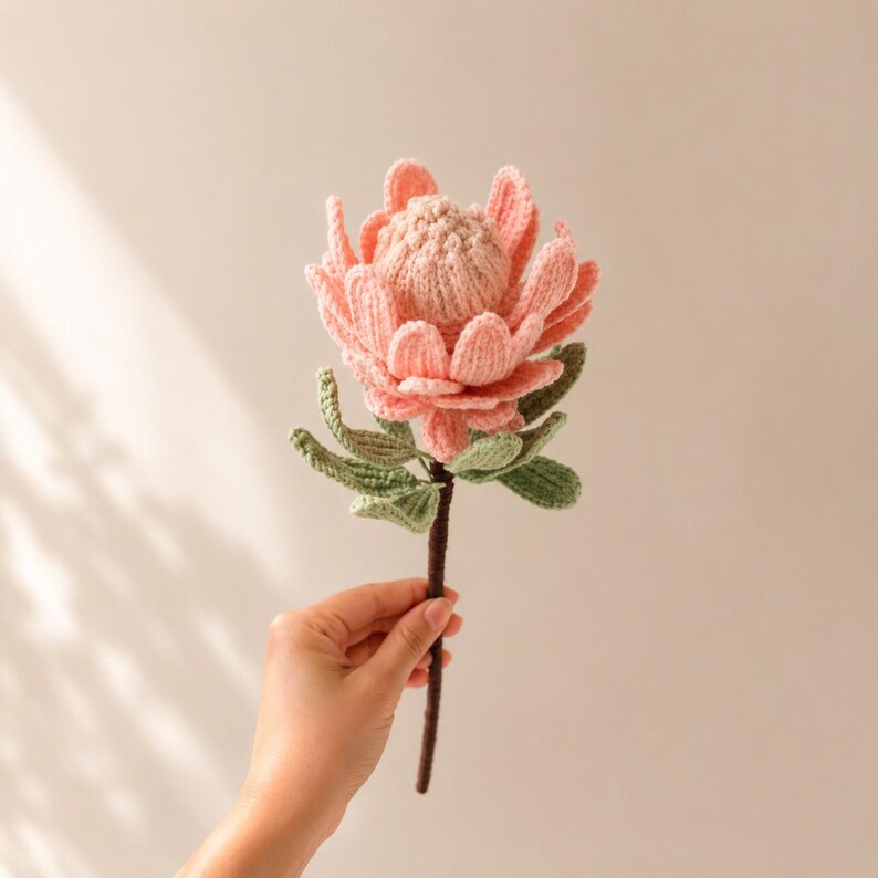 May include: A crocheted protea flower featuring soft pink petals with a textured, rounded center and several sage green leaves attached to a thin, dark brown stem. The handcrafted textile bloom mimics the appearance of a real king protea.