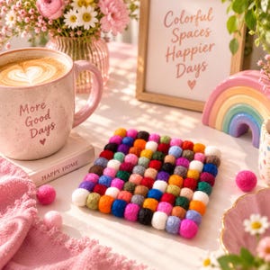 Puede incluir: Un posavasos cuadrado hecho de bolas de fieltro multicolores en tonos rosa, naranja, amarillo, verde, azul y morado. El posavasos está dispuesto en un patrón de cuadrícula y se encuentra sobre una superficie junto a una taza de cerámica con el texto More Good Days.