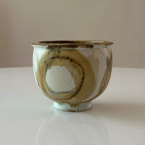 Peut inclure: Un bol en céramique fait à la main avec une base blanche et un motif en spirale marron et beige. Le bol a une forme arrondie et une petite base robuste. Le bord du bol est de couleur marron foncé.
