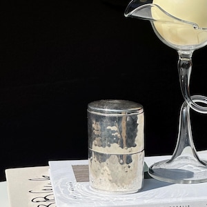 May include: A silver metal container with a hammered texture sits atop a stack of books. A clear glass candle holder with a large, round, cream-colored candle is in the background. The books have white covers with black text.