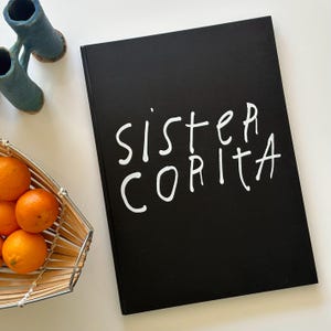 May include: A black book with the words "sister CORITA" in white handwritten font. The book is on a white surface, with a basket of oranges and a blue ceramic sculpture in the background.