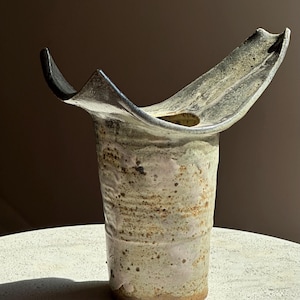 May include: A ceramic vase with a cylindrical body and a wide, flared, asymmetrical rim. The surface features a mottled, speckled glaze in shades of cream, beige, and light brown, with dark, charred-looking edges along the top rim opening.