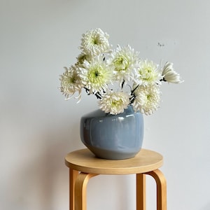 May include: A bouquet of white chrysanthemums in a blue ceramic vase on a wooden stool.