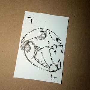 Puede incluir: Un boceto en blanco y negro de una calavera de gato con dientes afilados y un dise&ntilde;o detallado. El dibujo est&aacute; sobre un papel rectangular blanco con dos conjuntos de dos s&iacute;mbolos en forma de cruz encima y debajo de la calavera.