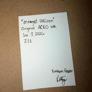 Puede incluir: Una tarjeta blanca con texto manuscrito que dice "enraged raccoon" Original ACEO ink Jan 3 2020 212. La tarjeta est&aacute; firmada "Kaithlynn Frazier" con una firma debajo.