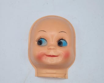 May include: A vintage doll head made of peach-colored plastic. The doll head has blue eyes, long eyelashes, and red cheeks and lips. The head is bald and has a small neck.