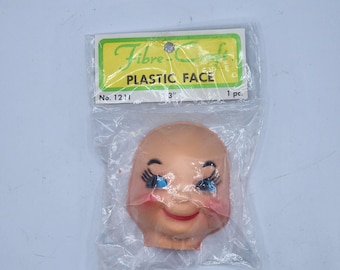 May include: A plastic doll face, packaged in a clear plastic bag. The face is peach-colored with blue eyes, black eyelashes, and a red smiling mouth. The package has a green and yellow label that reads "PLASTIC FACE" and "3" inches.