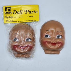 May include: Two vintage doll heads with brown skin, rosy cheeks, and red lips. One head is in a plastic bag, and the other is loose. A yellow label reads "Doll Parts" by Yaley, "Crafting Supplies."