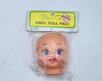 May include: A vintage vinyl doll face in a clear plastic bag. The doll face is peach-colored with blue eyes, long eyelashes, and a red mouth. The packaging includes a green and yellow label that reads "Fibre-C VINYL DOLL FACE". The doll face measures 3 inches.