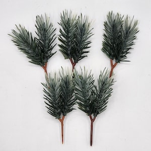 May include: Five artificial pine sprigs with brown stems. The sprigs are green with a slight white dusting.