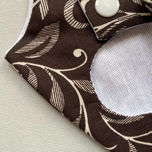 May include: A brown fabric with a white floral pattern and a white circle in the center. The fabric has a button on the top right corner.