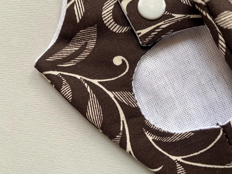 May include: A brown fabric with a white floral pattern and a white circle in the center. The fabric has a button on the top right corner.