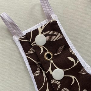 May include: A brown fabric with a white floral pattern and white snaps. The fabric is attached to white elastic straps.