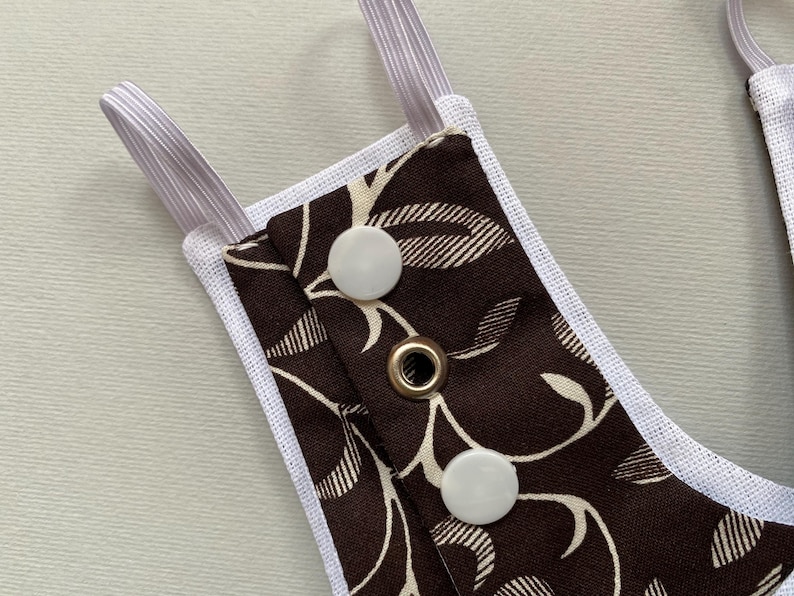 May include: A brown fabric with a white floral pattern and white snaps. The fabric is attached to white elastic straps.