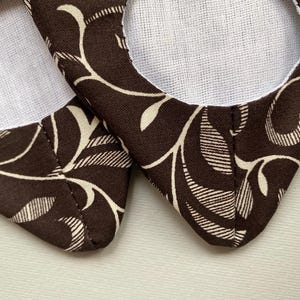 May include: A pair of brown fabric slippers with a white floral pattern. The slippers have a white lining and a pointed toe.