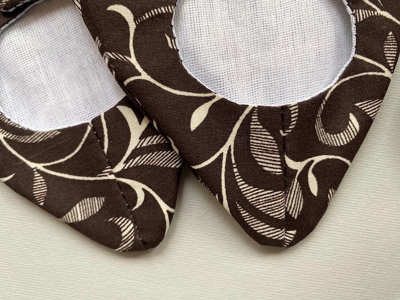 May include: A pair of brown fabric slippers with a white floral pattern. The slippers have a white lining and a pointed toe.