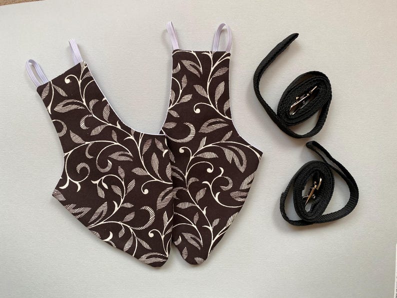 May include: Two brown and white patterned fabric pet harnesses with white straps and two black leashes with silver clasps.