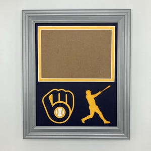 May include: A silver picture frame with a navy blue mat and a yellow and white baseball glove and player silhouette. The frame has a brown insert.