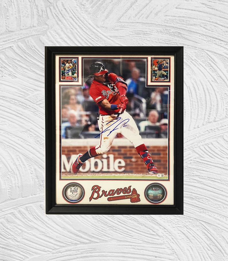 May include: A framed photo of a baseball player in a red and white uniform swinging a bat. The player is wearing a white helmet and has a blue signature on the photo. The photo is surrounded by smaller photos of other baseball players and has the words "Braves" and "Mobile" printed on it.