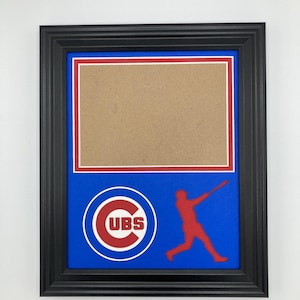 May include: A black rectangular frame containing a blue mat board with a tan rectangular opening at the top. The mat features a red and white Chicago Cubs logo on the left and a red silhouette of a baseball player swinging a bat on the right.