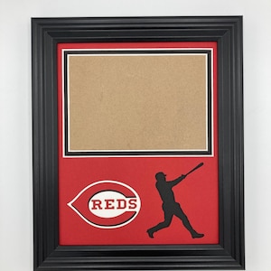 May include: Black framed picture with a red background featuring the Cincinnati Reds logo and a silhouette of a baseball player swinging a bat. The frame has a white and black border around the picture.