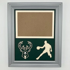 May include: A silver picture frame with a green mat and a white silhouette of a basketball player and the Milwaukee Bucks logo.