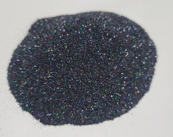 May include: A pile of dark glitter with iridescent flecks. The glitter appears black with hints of rainbow colors. The glitter is spread out on a white surface.