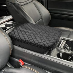May include: Black, quilted leather car console cover. The cover is diamond-stitched and fits over the center console of a vehicle. The interior of the car is black leather with silver accents.