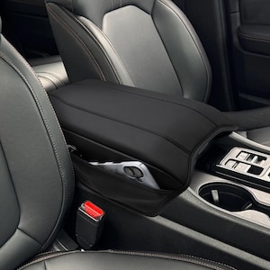 May include: Black car armrest cover with a built-in pocket. The cover is made of a smooth, leather-like material and is designed to fit over the center console armrest. A smartphone is visible in the pocket. The car seats are black.