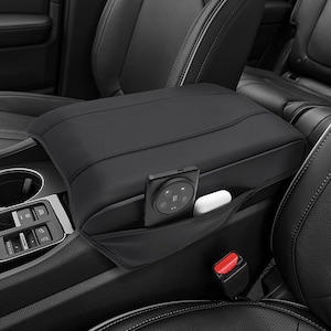 May include: Black car armrest cover with a pocket, holding a smartphone and wireless earbuds. The cover is installed in a car, with black leather seats and a red seatbelt buckle visible.