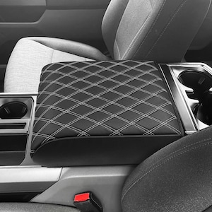 May include: Black car armrest with a diamond-stitched pattern and white thread. The armrest is rectangular and appears to be made of leather or a similar material. It is installed in a car, with the seats and console visible.
