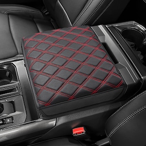 May include: Black car armrest cover with red diamond stitching. The cover is made of a smooth, leather-like material and is designed to fit over the center console armrest. The red stitching provides a contrasting accent to the black cover.