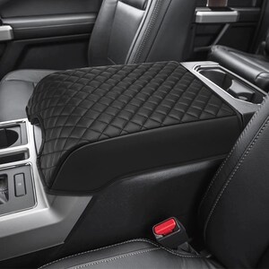 May include: Black quilted leather center console armrest cover for a car. The cover has a diamond pattern and is designed to fit over the existing armrest.