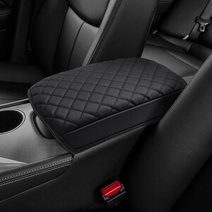 May include: Black car armrest with a quilted diamond pattern. The armrest is made of a smooth, leather-like material and is positioned between the front seats of a vehicle. The interior of the car is black.
