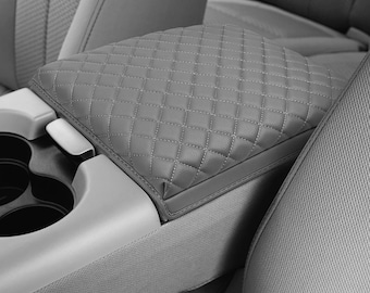 May include: Gray, quilted car armrest cover. The cover is diamond-stitched and fits over the center console armrest. The cover is shown in a car interior with cup holders and seats.