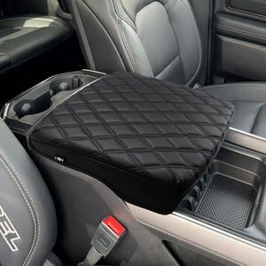 May include: Black quilted leather center console armrest cover for a car. The cover has a diamond pattern and a small logo on the side.