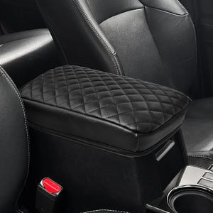 May include: Black quilted leather armrest cover for a car. The cover is designed to fit over the center console armrest and provides a more comfortable and stylish look.