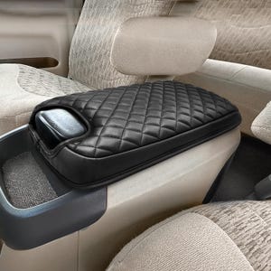 May include: Black quilted car armrest cover. The cover is diamond-stitched and fits over the center console. The interior of the car has beige seats and a headrest.