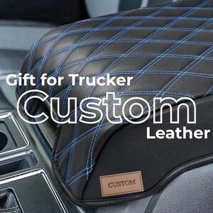 May include: A black leather vehicle centre console cover featuring a diamond-stitched pattern with blue thread. The cover includes a small rectangular brown patch with the word CUSTOM embossed on it. Text overlay reads Gift for Trucker Custom Leather.