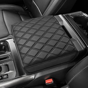 May include: Black, diamond-stitched car armrest cover. The cover is made of a smooth, leather-like material and is designed to fit over the center console armrest. The stitching creates a quilted pattern.