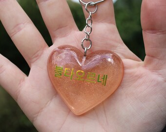 Puede incluir: Un llavero en forma de corazón rosa con purpurina dorada y el texto "말티요트니" en letras doradas.