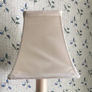 May include: A white lampshade with a square shape and a slightly curved bottom edge. The lampshade is made of a fabric material and has a smooth, even texture.
